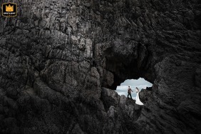 In Fujian, China, the bride and groom pose for a seaside portrait, framed through a natural opening in a rock formation resembling a cave, emphasizing the dramatic coastal landscape and their elegant presence against the ocean backdrop.